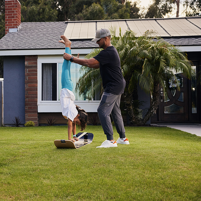 Man assisting a child do a handstand on a slackboard in a front yard.
