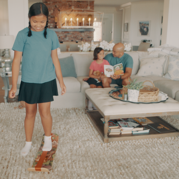 Girl balancing on GIBBY SlackBoard, while her dad reads GIBBY book to her sister on a couch in the background